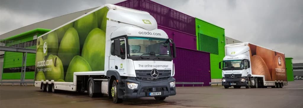Ocado Mercedes tractor units with double-deck trailers in apple and wood liveries at a fulfilment centre