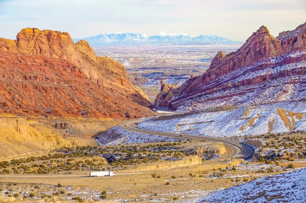 A white articulated lorry on a highway through red-rock desert, with snow on the slopes and mountains on the horizon