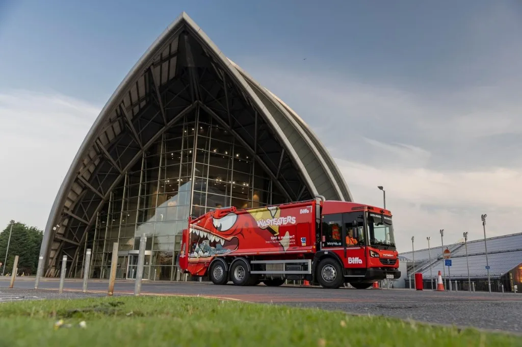 Red Biffa refuse collection vehicle with a cartoon monster on the side, parked in front of a glass building