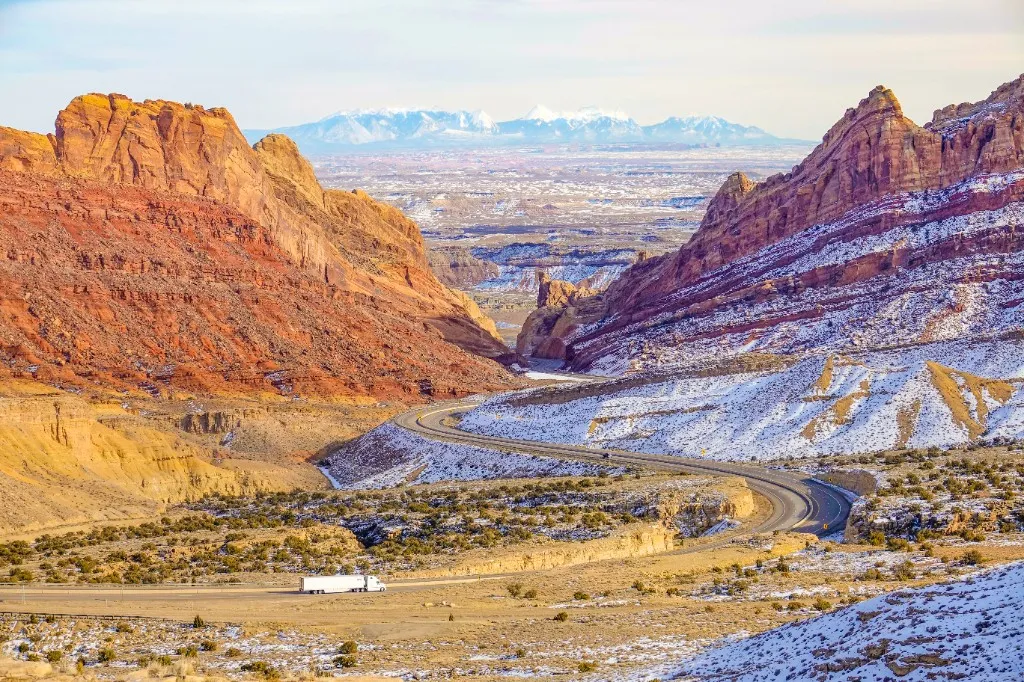 A white articulated lorry on a highway through red-rock desert, with snow on the slopes and mountains on the horizon