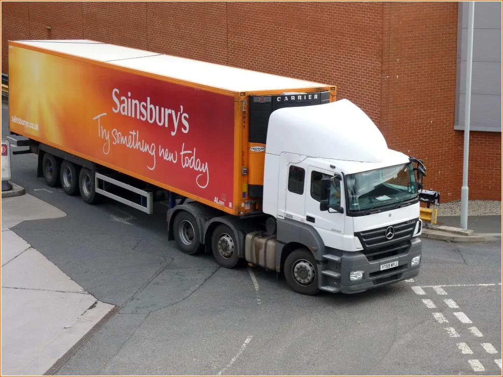 White Mercedes tractor unit pulling a Sainsbury’s refrigerated trailer in an orange and red livery, on an industrial estate