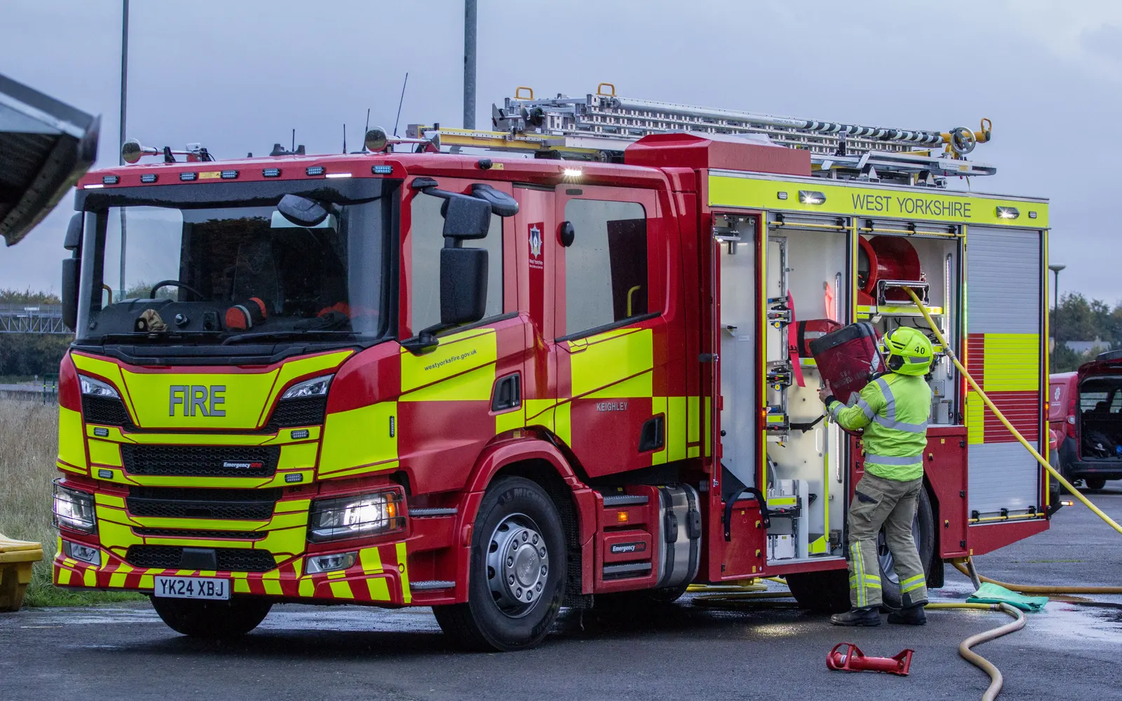 West Yorkshire Fire and Rescue Service Scania pump appliance in red and yellow livery, with a firefighter at the pump bay — photo by Graham Mitchell, CC BY 2.0